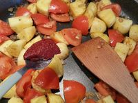 2 - Dans un grand plat de cuisson (style plat à paella, wok ou tajine), faire revenir les merguez dans un peu d'huile d'olive jusqu'à légère coloration. Retirer et réserver. Dans le même plat faire suer l'oignon et l'ail 2 à 3 mn, rajouter les tomates, mélanger et laisser cuire quelques minutes, puis rajouter les pommes de terre, mélanger. Incorporer successivement, l'harissa, le ras el hanout, le paprika, le curcuma, le concentré de tomates, le piment vert et le persil (ou la coriandre - en réserver un peu pour la présentation), saler et poivrer.