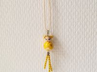 La petite kokeshi prénommée Hikari est montée sur un collier en cordon de coton écru de 56 cm de tour de cou. La poupée mesure 2.5 cm et est agrémentée de perles de rocaille jaune et dorée