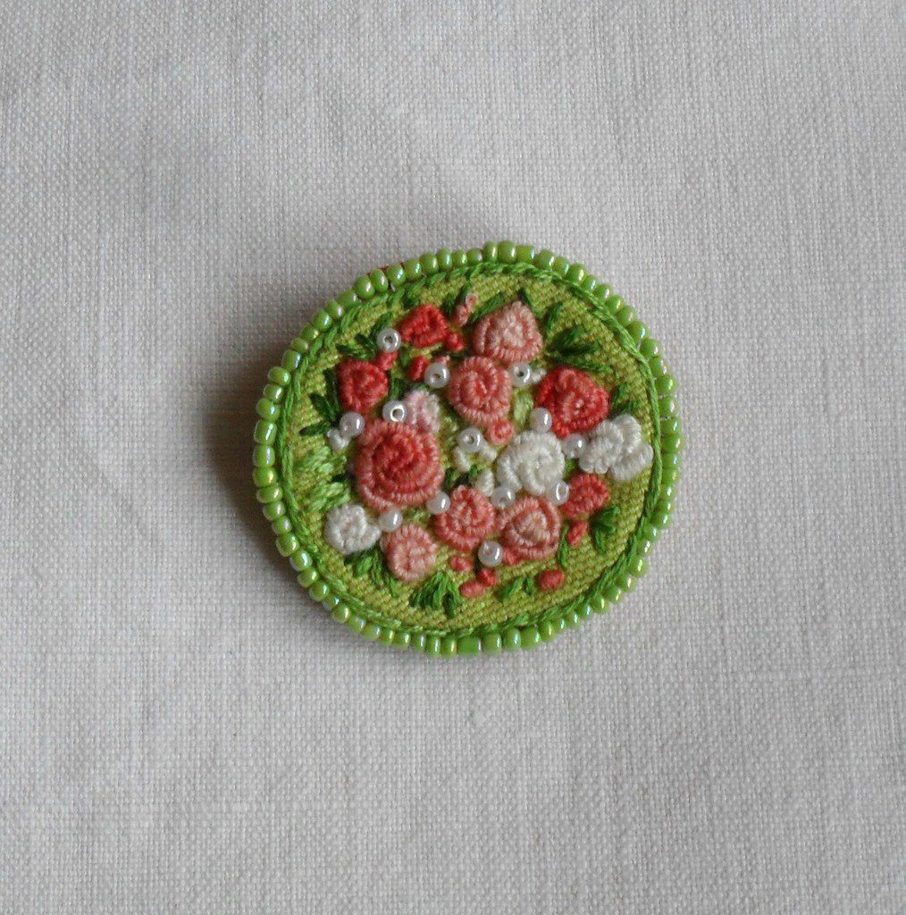 Broche fantaisie , de forme ronde, au centre des roses brodées main sur fond vert dans un dégradé de couleur allant du blanc au saumon, ajout de perles blanches nacrées, dos en cuir coté daim ton orangé, entourage brodé et rang de perles nacrées vert pale, attache de couleur bronze diamètre 4.7 cm