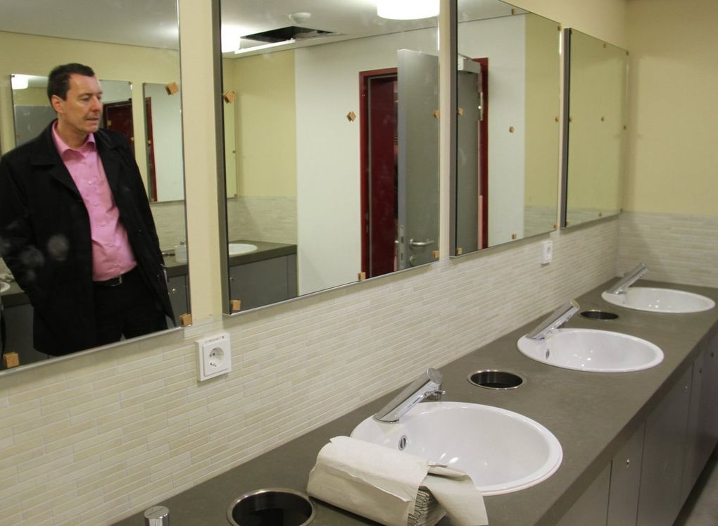 Im Untergeschoss untergebracht sind die Technik, Lager- und Nebenräume, Sanitärtrakt und außen auch ein öffentliches WC.  Kurz vor den Toiletten abgetrennt wurde ein fester Garderobenraum. Bei kleineren Veranstaltungen sind aber auch oben mobile Garderobenelemente vorgesehen. Auf der nördlichen Foyerseite im UG ermöglicht eine Rampe einen barrierefreien Zugang von der Straße aus. Nach oben gelangt man von hier über einen Personenaufzug.  Im Bild überzeugt sich der Bürgermeister von der Funktionsfähigkeit des neuen Behindertenaufzuges. Erhalten blieb im Bereich des Treppenabgangs vom Foyer zum Untergeschoss der den Main symbolisierende Glasschmuck in der Backsteinwand, die auf der anderen Seite zum Foyer hin wie die Wände im großen Saal verkleidet wurde.  Die aufgelassene Kegelbahn bietet nun ausreichend Lagerflächen. Imposant ist die Technik nebenan im Heizungsraum, in dem auch ein Blockheizkraftwerk installiert werde.