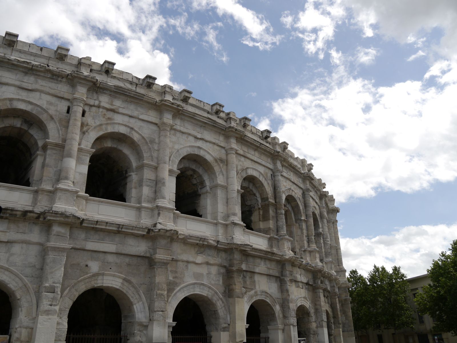 La façade extérieure des arènes de Nîmes se détache sur un ciel bleu légèrement nuageux.