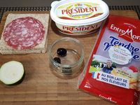 1 - Couper la grande rondelle de saucisson en deux. Tailler dans la rondelle de courgette 4 petits disques avec un petit emporte-pièce rond. Découper les olives dénoyautées en rondelles fines à déposer sur les disques de courgette. Trancher le pain de mie en deux dans le sens de la diagonale. Beurrer le pain de mie. déposer dessus la demi-tranche de saucisson comme indiqué sur la photo. Placer dessus des rectangles de fromage et recouvrir des triangles de pain de mie restants.