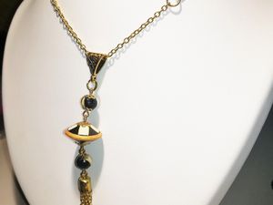 Le Collier est composé :  - d'une chaîne en métal doré de 65 cm de tour de cou,  - d'un bijou composé de : - 2 perles en bois (diam. 0.60cm), d'une soucoupe en bois (diam. 2cm) peintes de couleur noir et or, - 1 floche en métal doré.